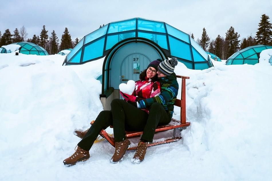 Aurelia Teslaru and Dan Moldovan sitting on a wooden sled in front of a glass igloo during winter at Kakslauttanen Arctic Resort Lapland